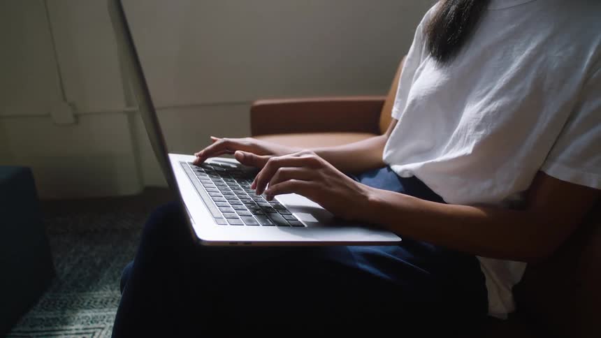 Woman wearing a white t shirt and blue pants sits in chair typing on a grey laptop next to an empty chair with a wall in the background and carpet on the floor