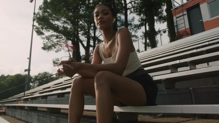 A woman with earbuds sitting on bleachers with trees the sky and clouds in the background