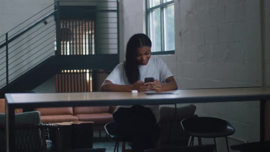 A young woman sits at a large table looking at her cell phone and smiling