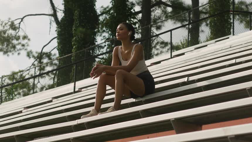 A woman wearing sneakers shorts and a tank top sits on bleachers outside in front of trees