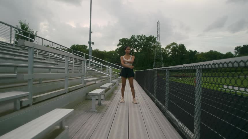 A young woman stretches her arms while standing in the bleachers beside an athletics track on a cloudy day