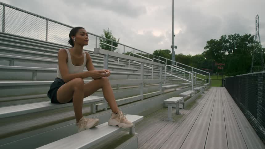 Woman sits on bleachers outside and readjust her headphones in the distance there are trees and grass under a grey cloudy sky