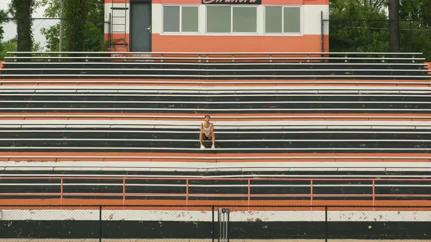 Woman wearing a tank top and shorts stands on black white and orange bleachers then sits down in the background is a building with a ladder and trees moving in the wind