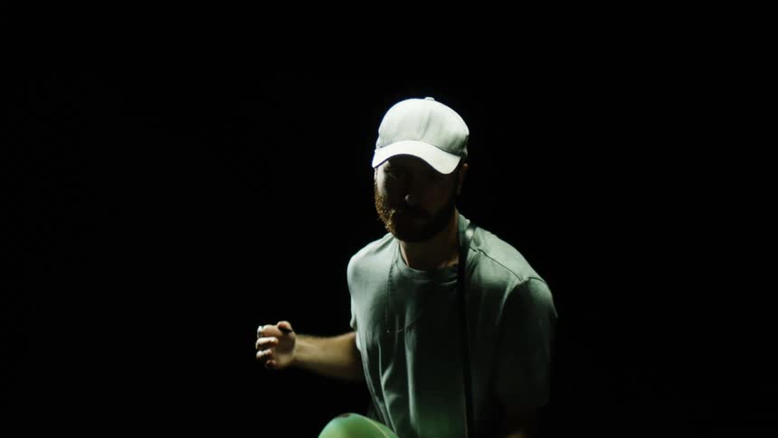 A man with a beard and wearing a baseball hat and t shirt strums on a guitar in slow motion in shadows with a black background