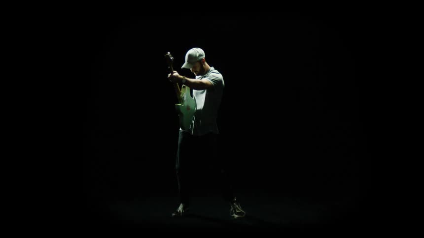 A man swings his guitar across his body as he strums it under a spotlight in a dark room