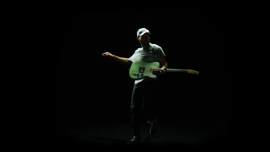 A guitarist swings his guitar exaggeratedly while playing it in a dark studio