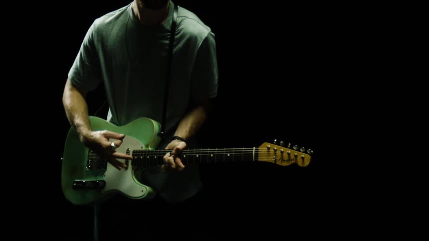 A man strums a bluegreen guitar under a light in the darkness