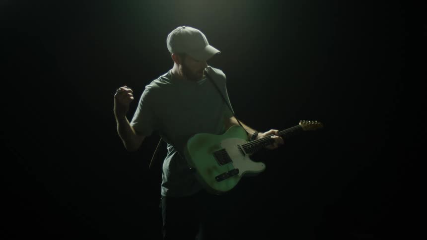 Man wearing hat t shirt and pants in the dark with spotlight on him plays an electric guitar