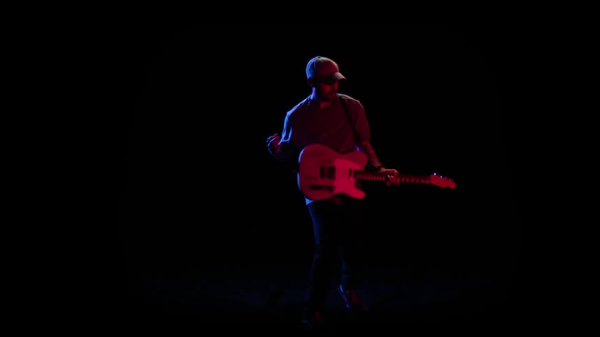 Man with a beard wearing a hat shirt pants and shoes plays a guitar with a pick in the dark