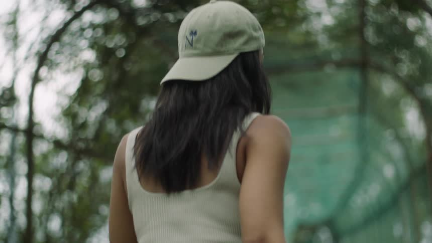 A young woman walking along a fenced walkway covered in vegetation turns to briefly smile into the camera before continuing on her way