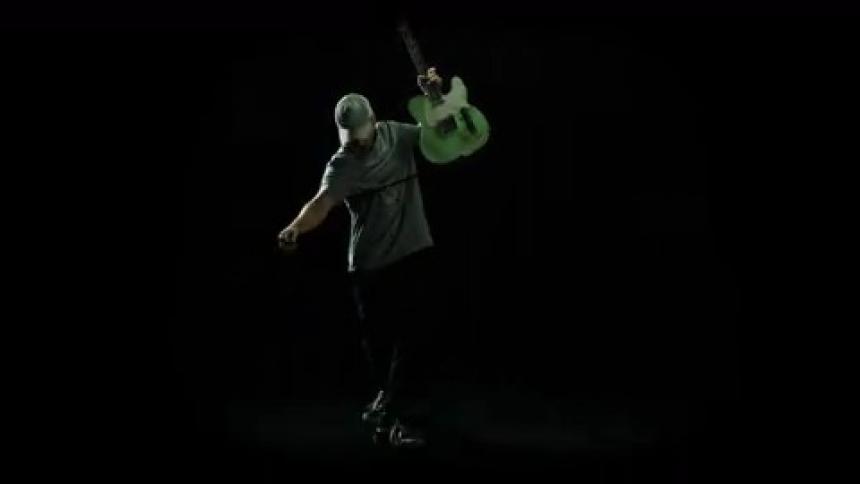 A man swings his guitar wide while standing under a flashing light in a dark place
