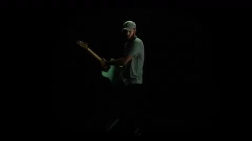 A man strums on a guitar as his body turns in slow motion on stage with a black background and brief strobes of light
