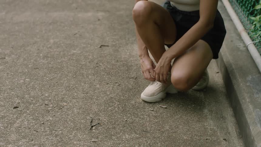 Woman wearing shorts crouches down near a chain link fence to tie her shoe laces