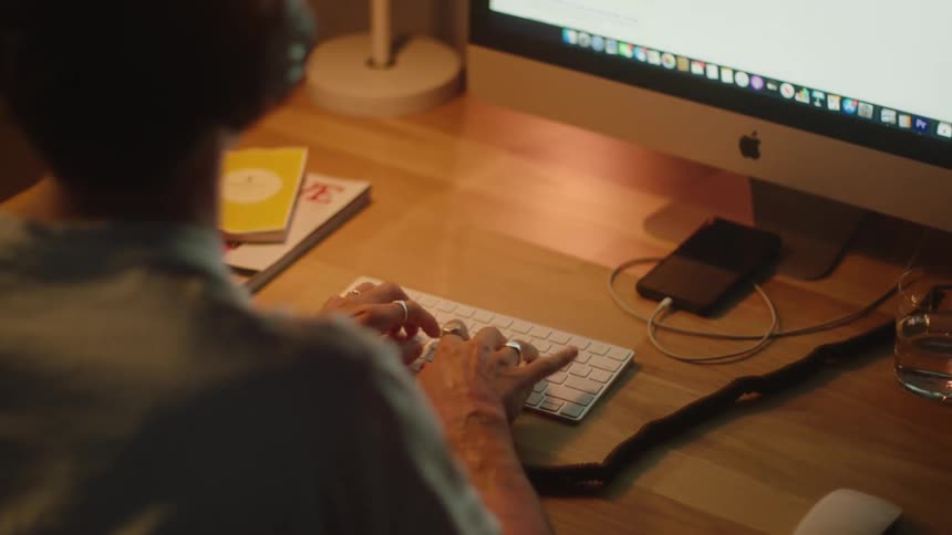 A man is wearing headphones while typing on his mac at a wooden desk