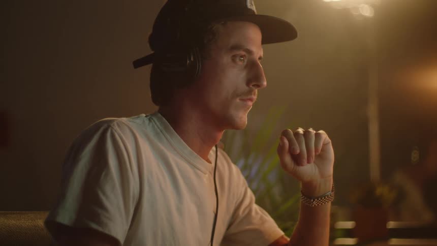 Man with a cap headphones and a white t shirt works on computer in a dark room