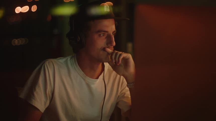 A man wearing headphones uses a computer at a desk indoors at night