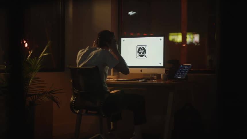 A male enters the room and sits down at a computer desk and puts headphones on his head