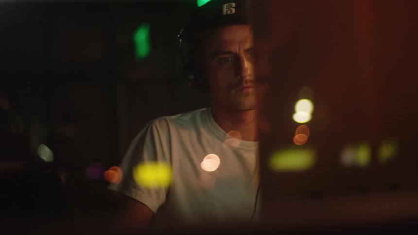 A man sits at a desk with a keyboard in a dim lit room at night with a contemplative focused face