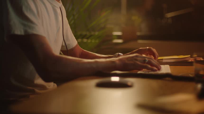 Person wearing white t shirt sits at table manipulating mouse and typing on keyboard
