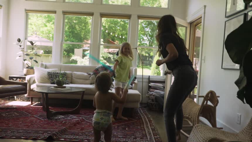 A mother and her two young daughters play with toy light sabers in the living room