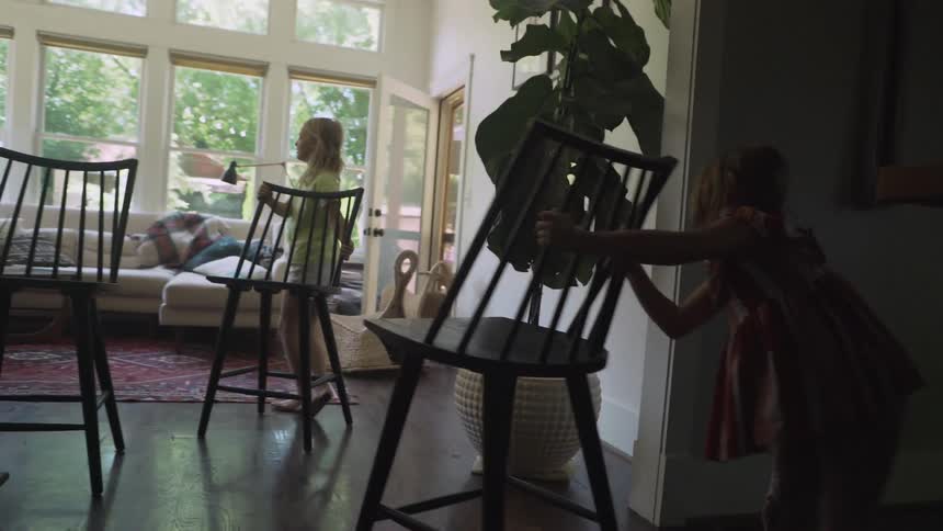A young girl pushes a chair through a house to the living room where her sisters await with their own chairs