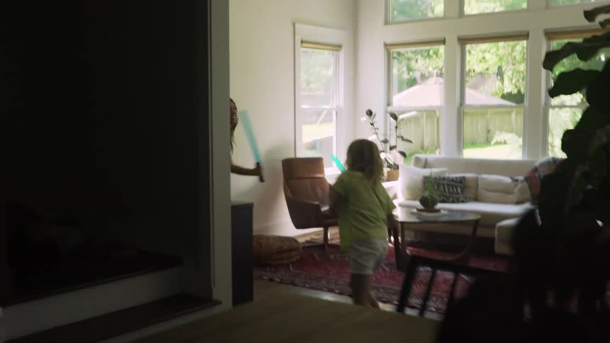 Mother and daughter play sword fight with light sabers in living room in front of a dinning room table and chairs behind them are a couch a chair a coffee table some plants pillows a throw blankets and windows through the windows you can see the back yard