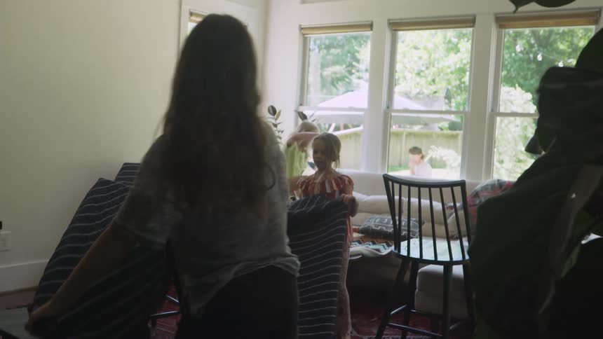 Kids stretch blanket over chairs in living room while mother sits under umbrella in yard