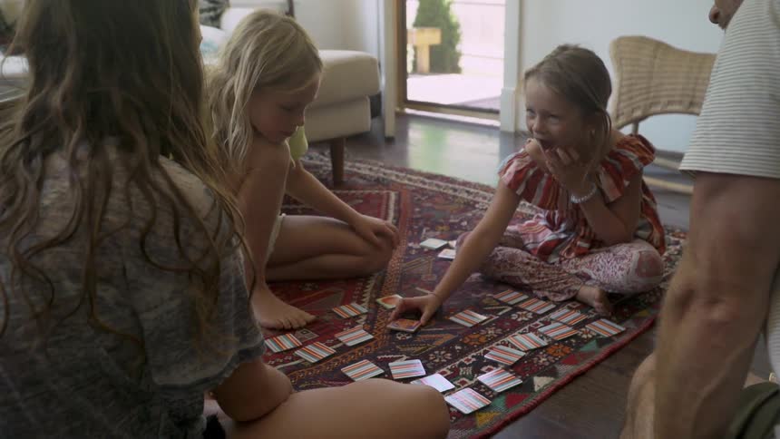 Two young girls sit on the floor playing a card game while their parents watch
