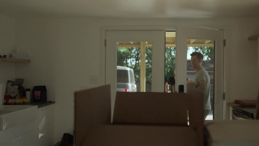 A husband and wife enter a home where there are open cardboard boxes on the counter and they bring in more boxes