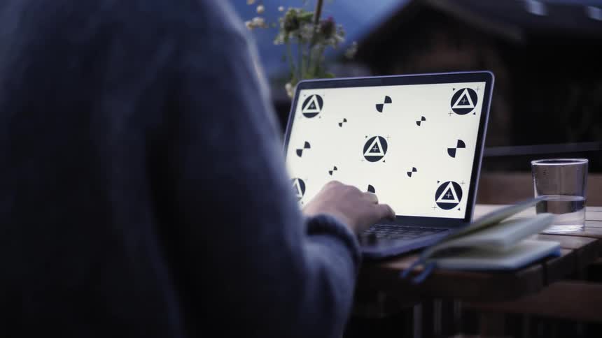 Person with a long sleeved shirt sits at a wooden table with a laptop book and glass of water typing at night beside flowers and in front of a house