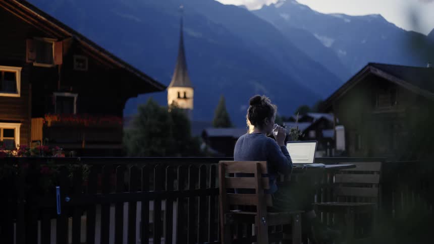 A woman sits at a small outside table with a laptop writing on something