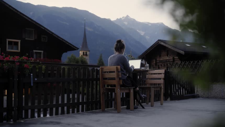 Young woman working on a laptop sitting at a table outdoors in a small town next to the mountains