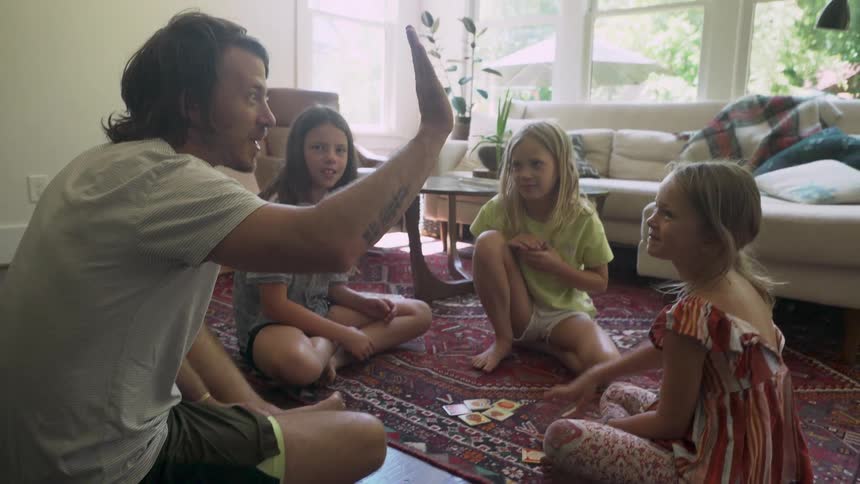 A man sitting on the floor playing cards with a group of girls high fives the girl sitting closest to him
