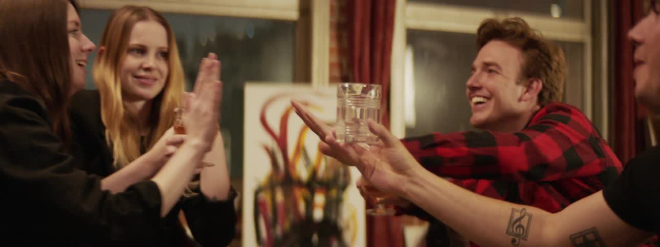 Two women and two men in long sleeves make a toast and high five at night sitting in front of a picture and some windows with red curtains
