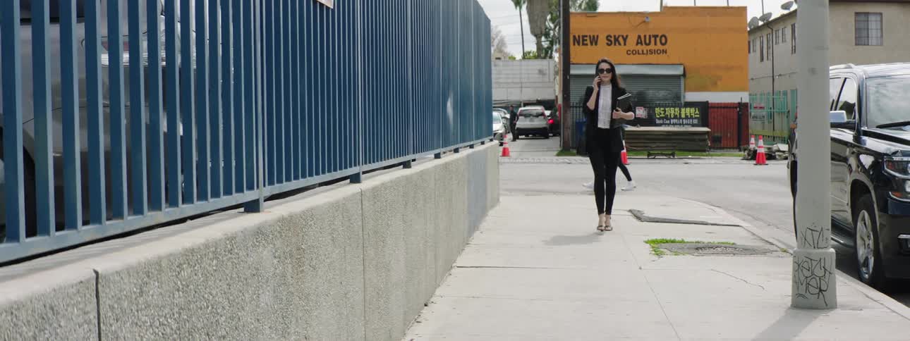 A woman in a suit and sunglasses talks on her smartphone while walking along a city sidewalk carrying a binder