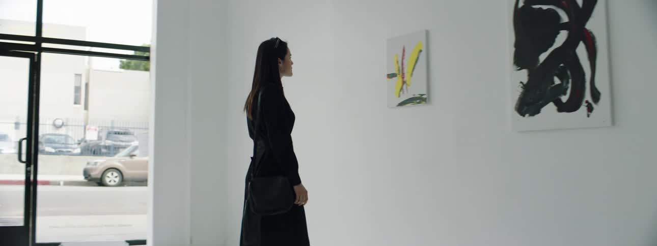 A woman in a black suit enters an art gallery and views art on the white walls