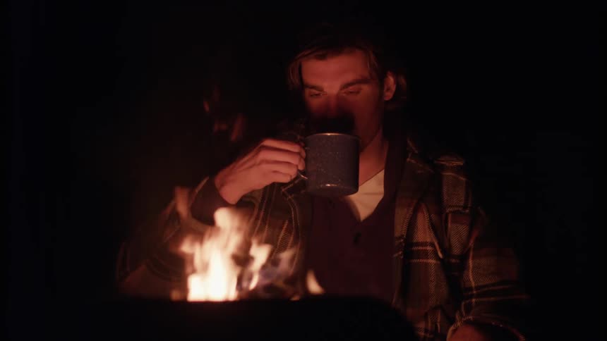 Man is sipping a drink from a mug while sitting outside by a fire at night