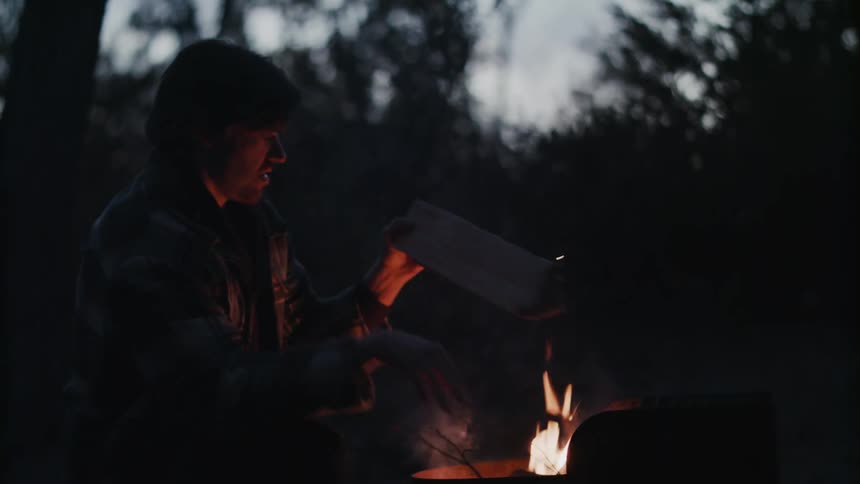 A man kneels before a fire burning in a barrel in a woodland and feeds it wood