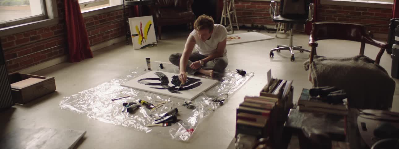 A man sits on the floor and uses a paintbrush to paint on a canvas