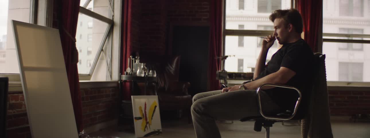 A man is sitting in an art studio holding a paint brush and staring at blank canvas