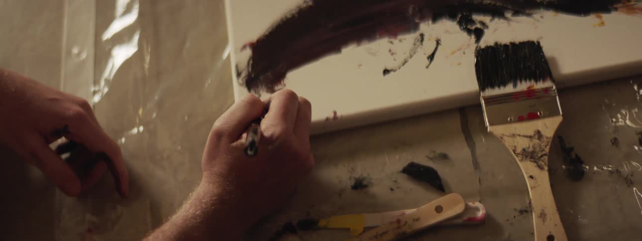 A hand is drawing on a white canvas that has black paint on it sitting on plastic and a paintbrush propped on the frame