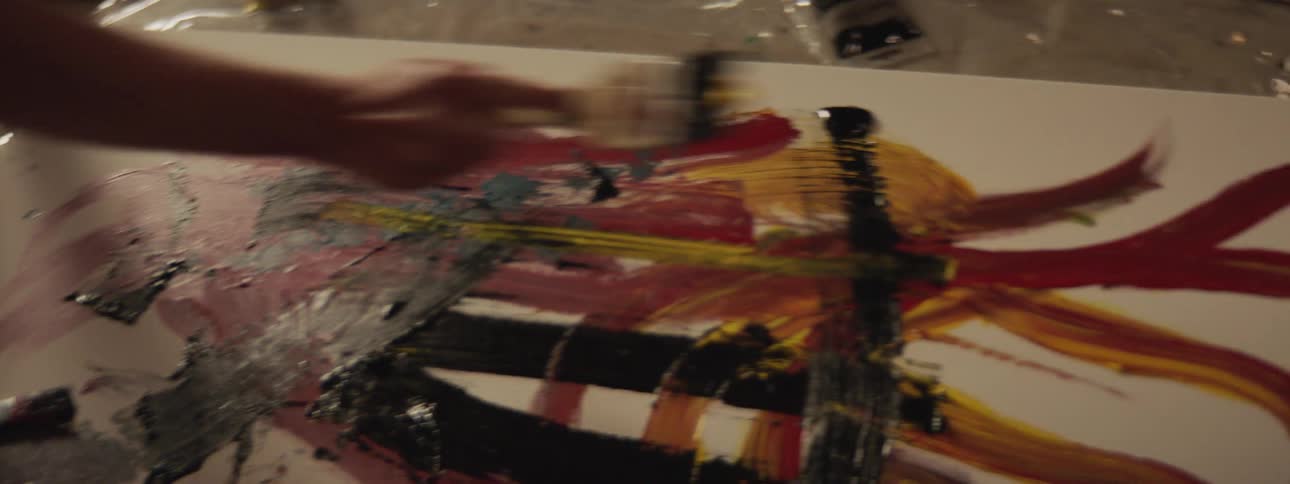 A man kneels on the floor using a paint brush to paint a canvas laying on the floor next to paint bottles in natural light