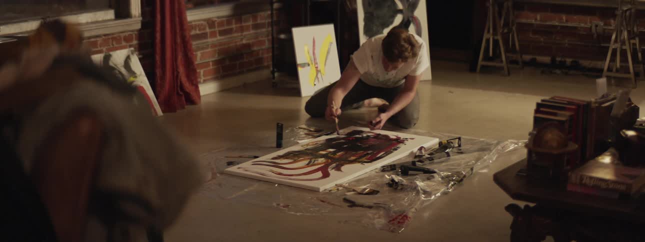 A man sits on a floor painting on a large canvas