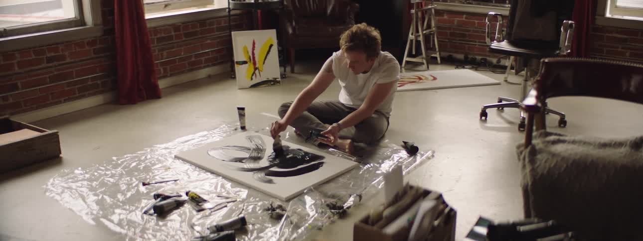 A man sitting in a room floor painting on a canvas with a brush