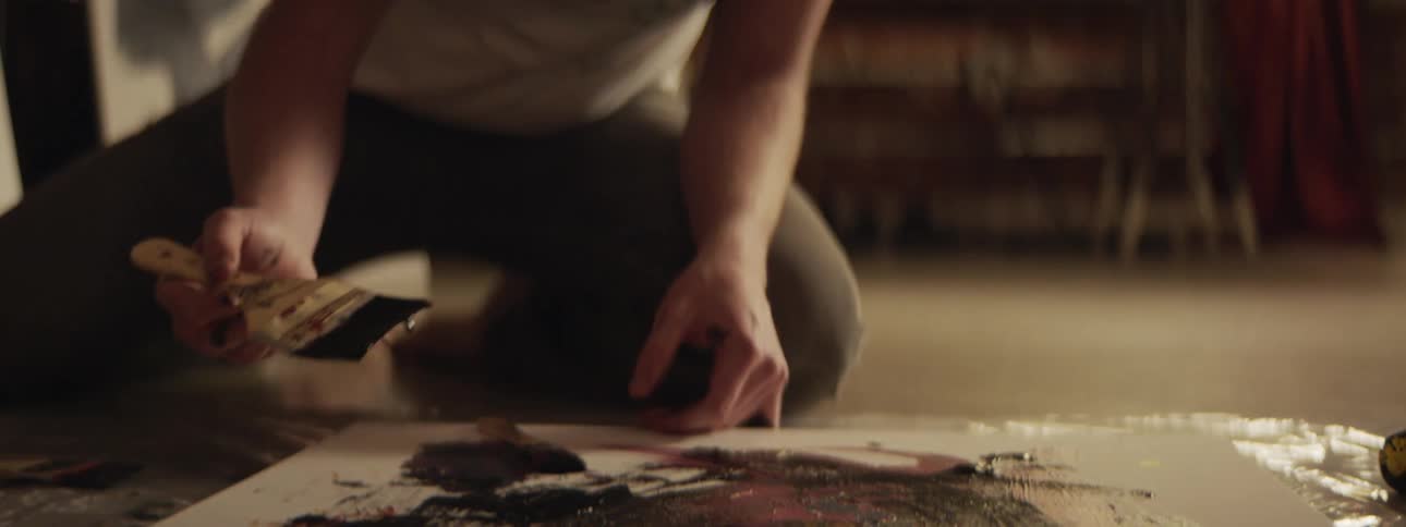 A man is kneeling on the floor indoors at night and painting on a canvas