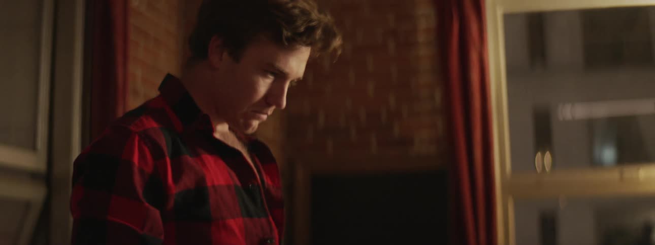 A man in a red and black flannel shirt places next to the window in his apartment at night