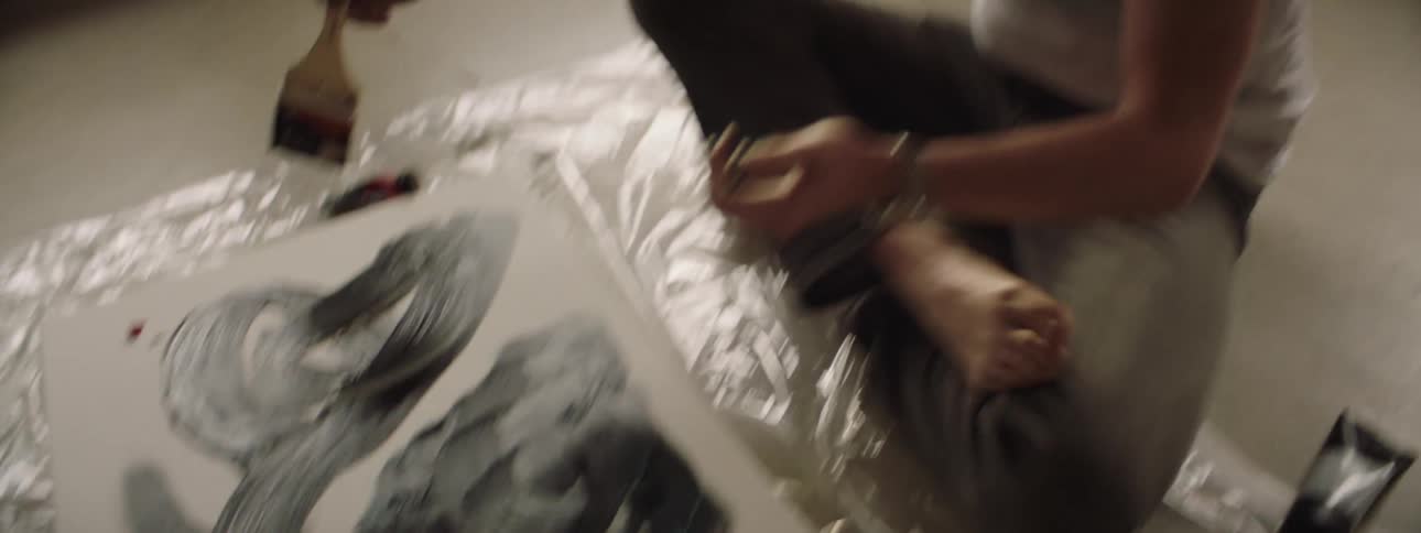 A man sits on the floor while painting on a canvas set on a plastic sheet