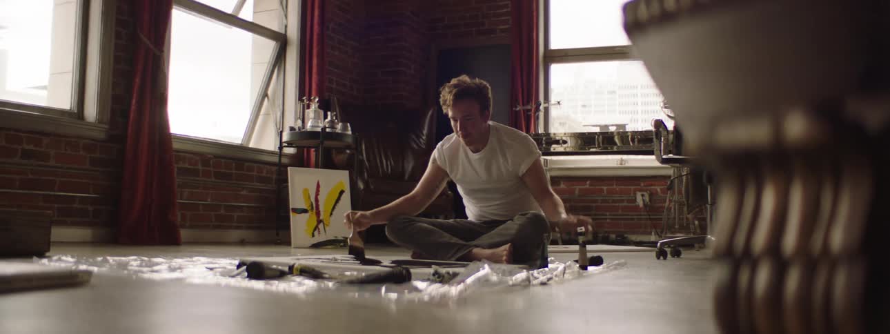 A caucasian man sitting on the floor cross legged painting on a canvas in a building