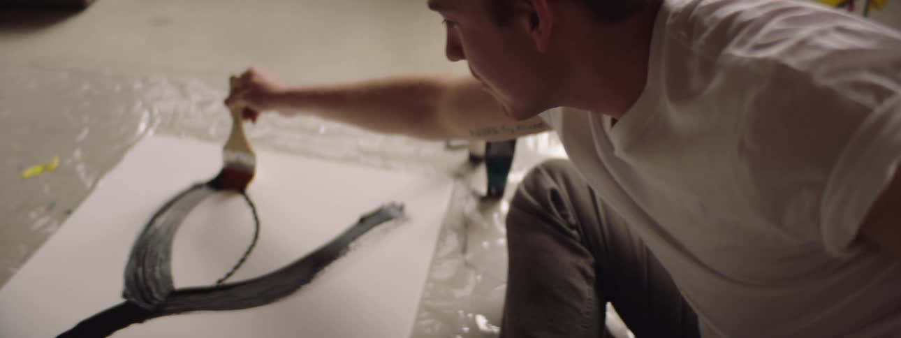 A man sitting on the floor with a cigarette in his mouth painting on a canvas with a paintbrush