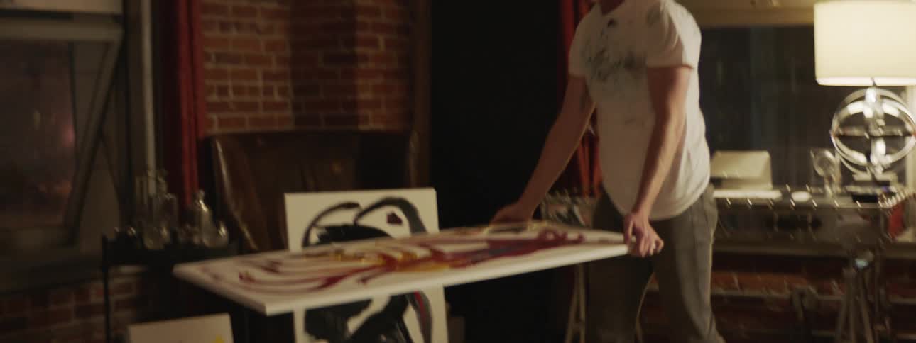 A man retrieves an unfinished painting from the floor to place it on a sheet of plastic as he prepares to paint